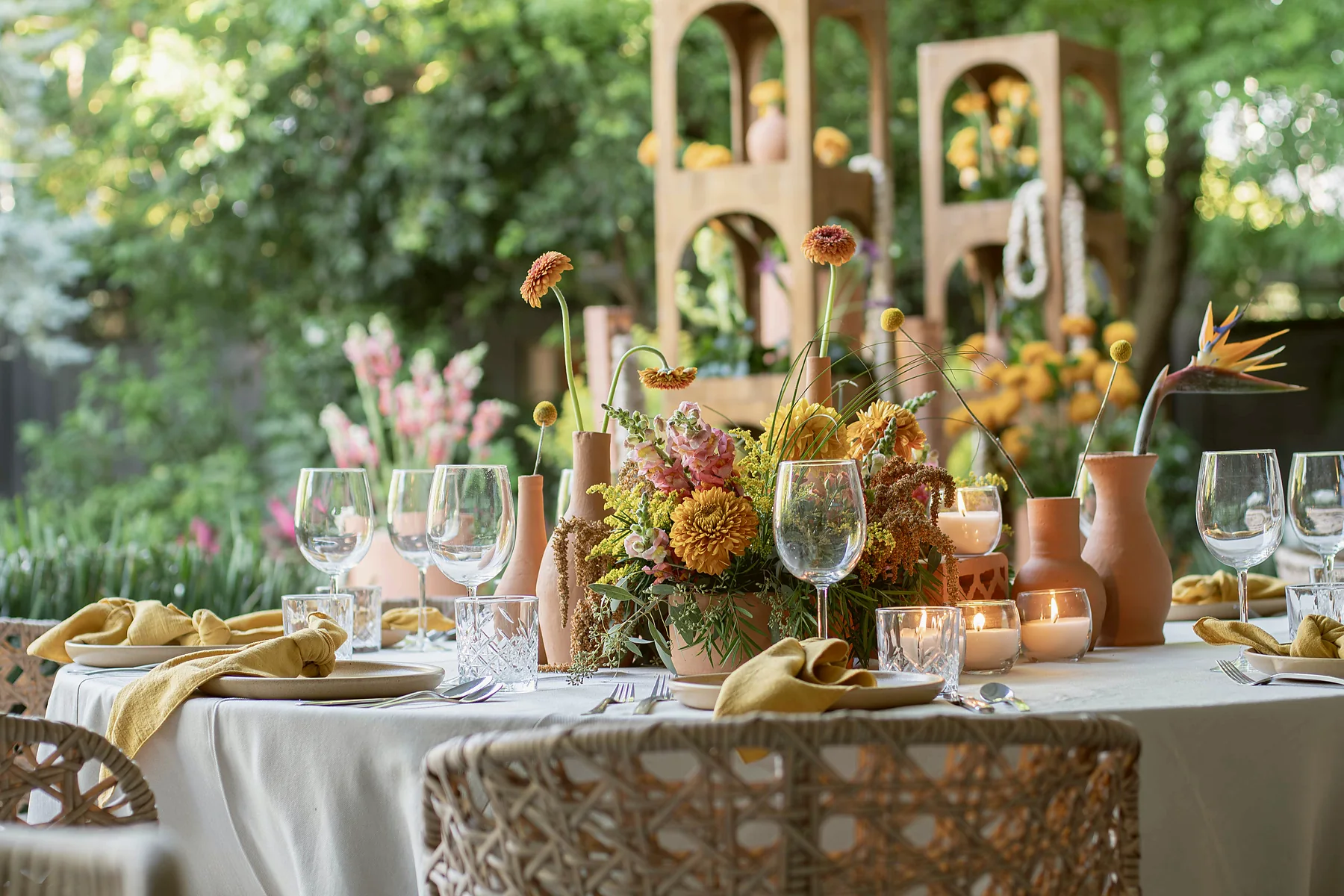 Mesa redonda montada con arreglo floral en tonos naranja y amarillo, con estructuras de madera con arcos al fondo del jardín